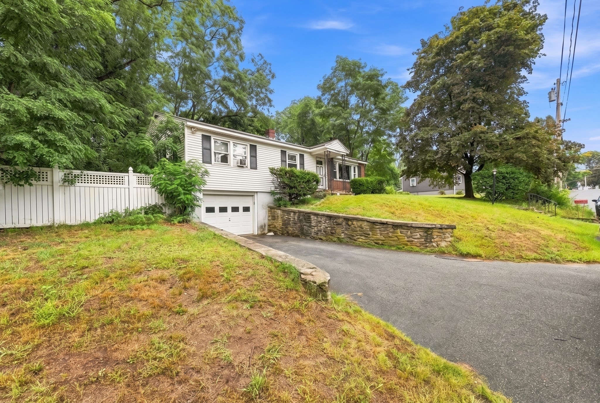 White single-story home with attached garage, sloped lawn, stone retaining wall, and mature trees.