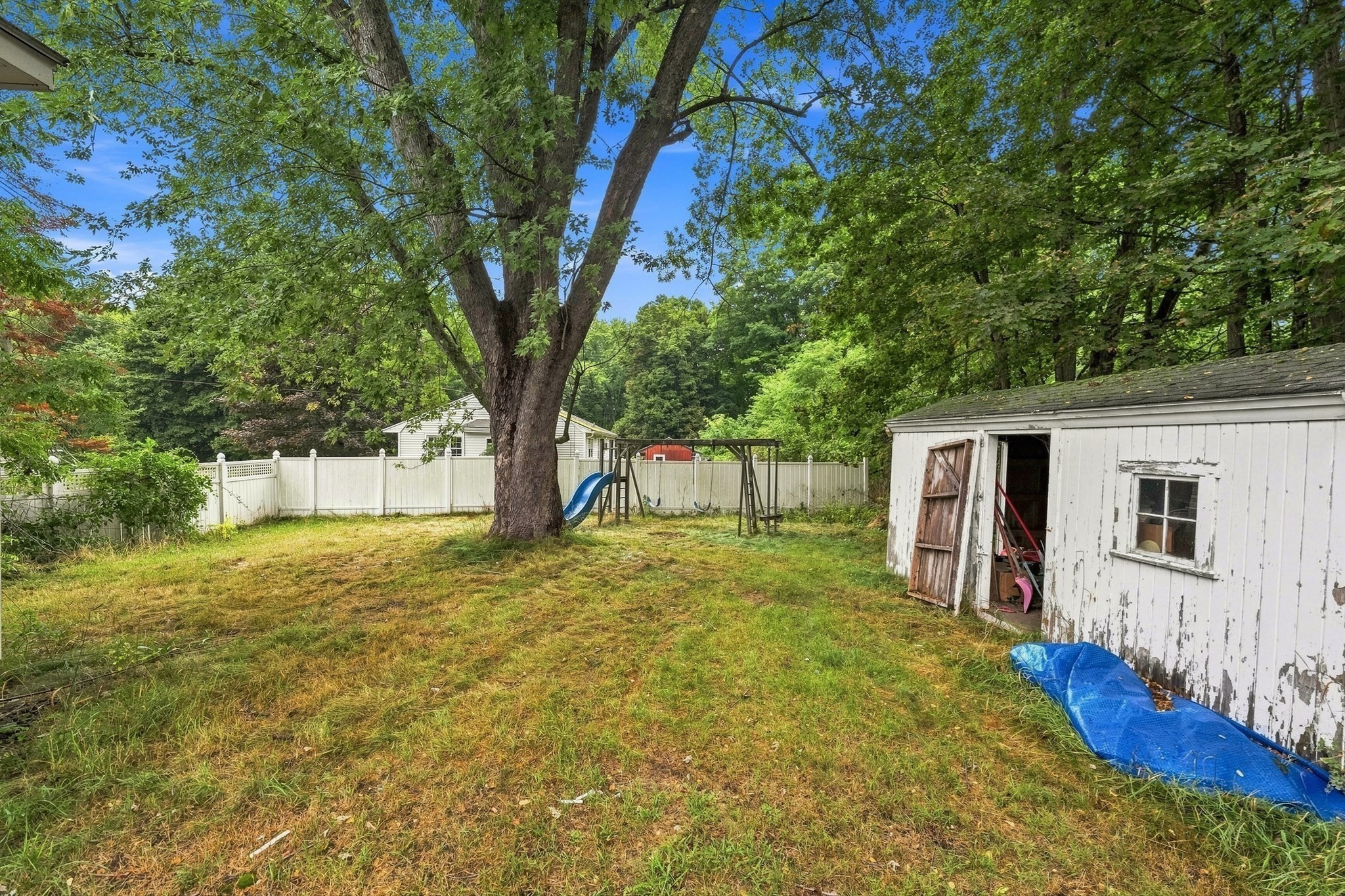 Backyard with a large shade tree, fenced swing set and slide, and a weathered white shed.