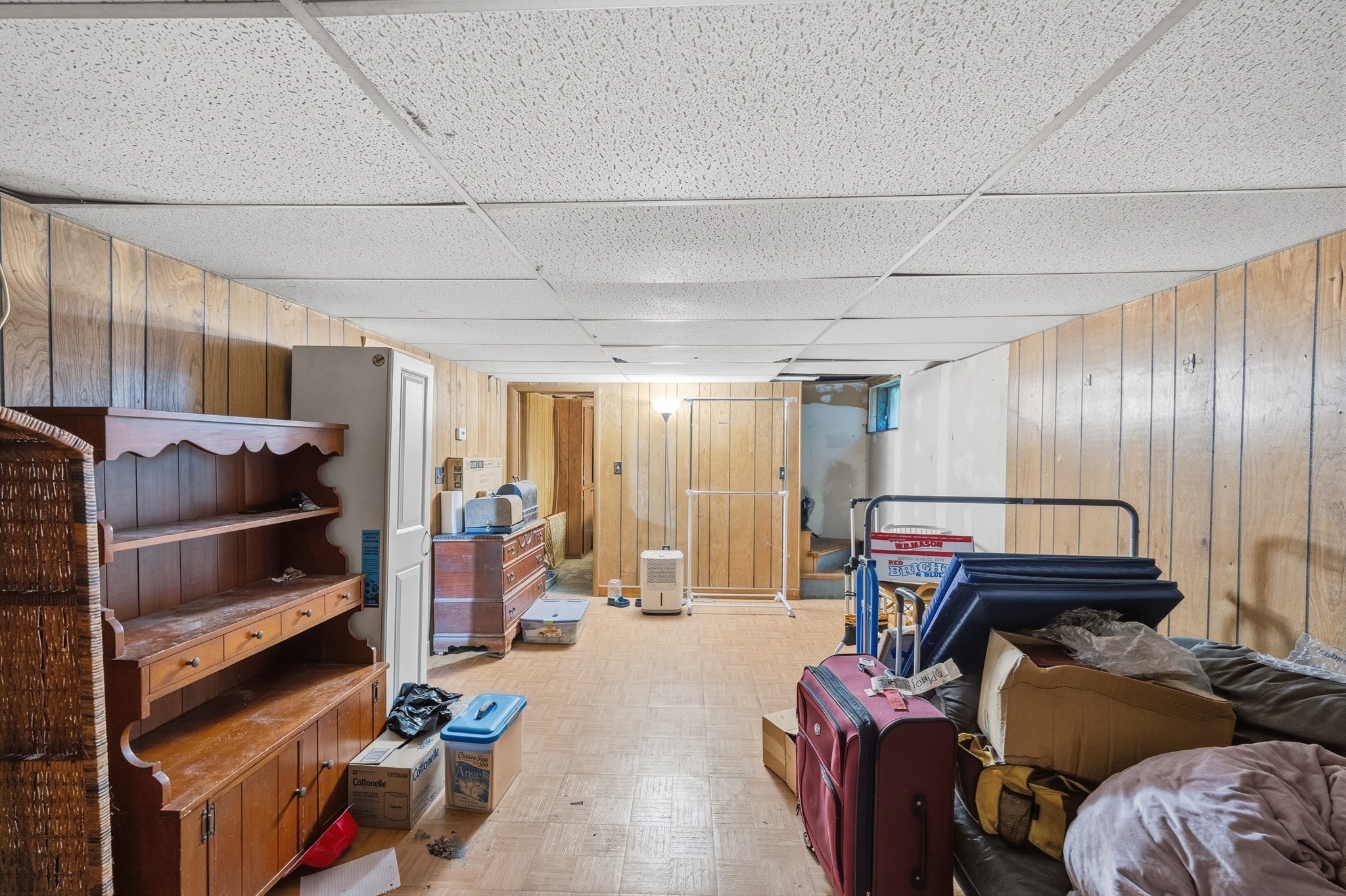 Basement storage room with wood-paneled walls, a drop ceiling, and belongings filling the space.