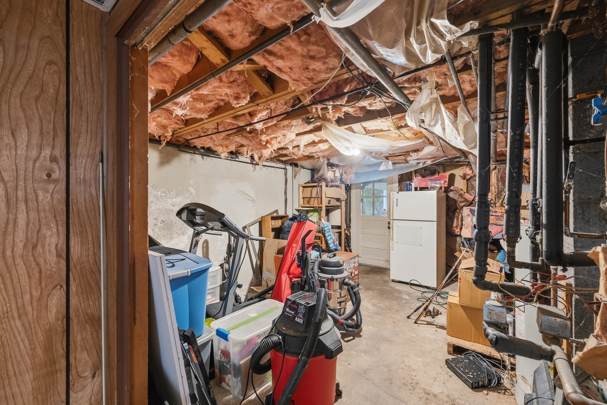 Basement storage area with exposed insulation, pipes, boxes, and a shop vacuum.