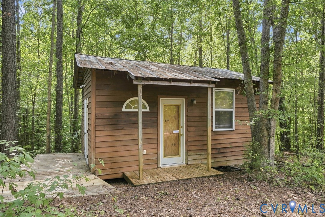 Small brown cabin with a metal roof and wooden deck nestled among leafy trees.