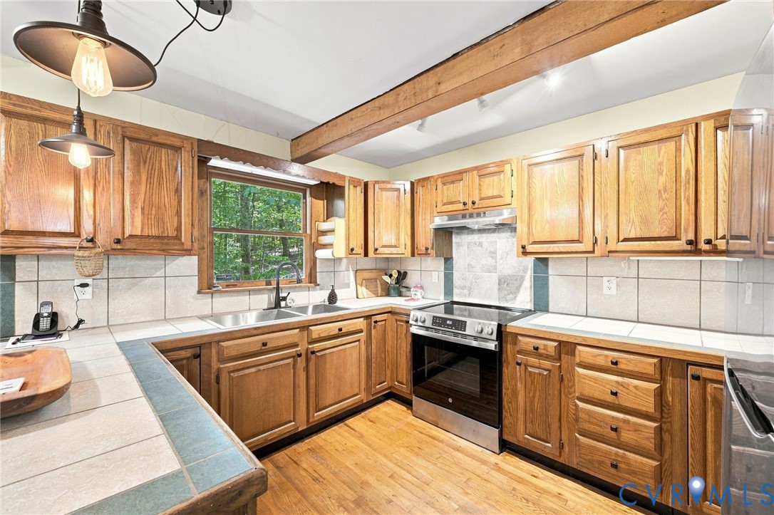 Kitchen with honey-toned wood cabinets, tiled counters, stainless-steel range, and a window overlooking trees.