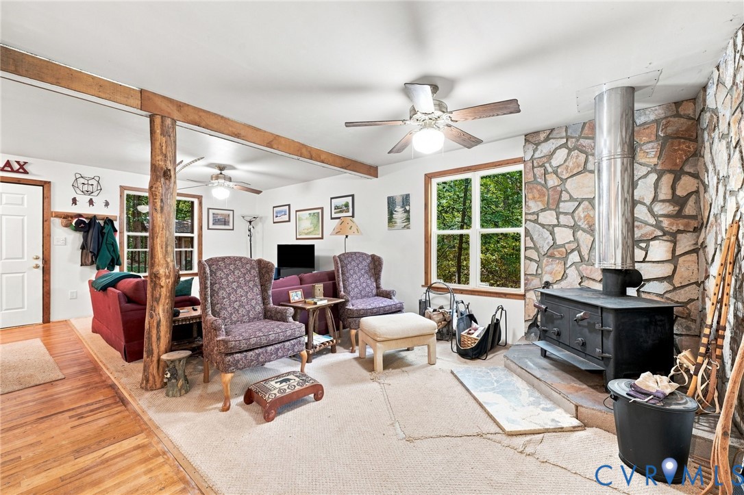 Living room with a stone fireplace wall, wood-burning stove, exposed beams, and windows overlooking trees.