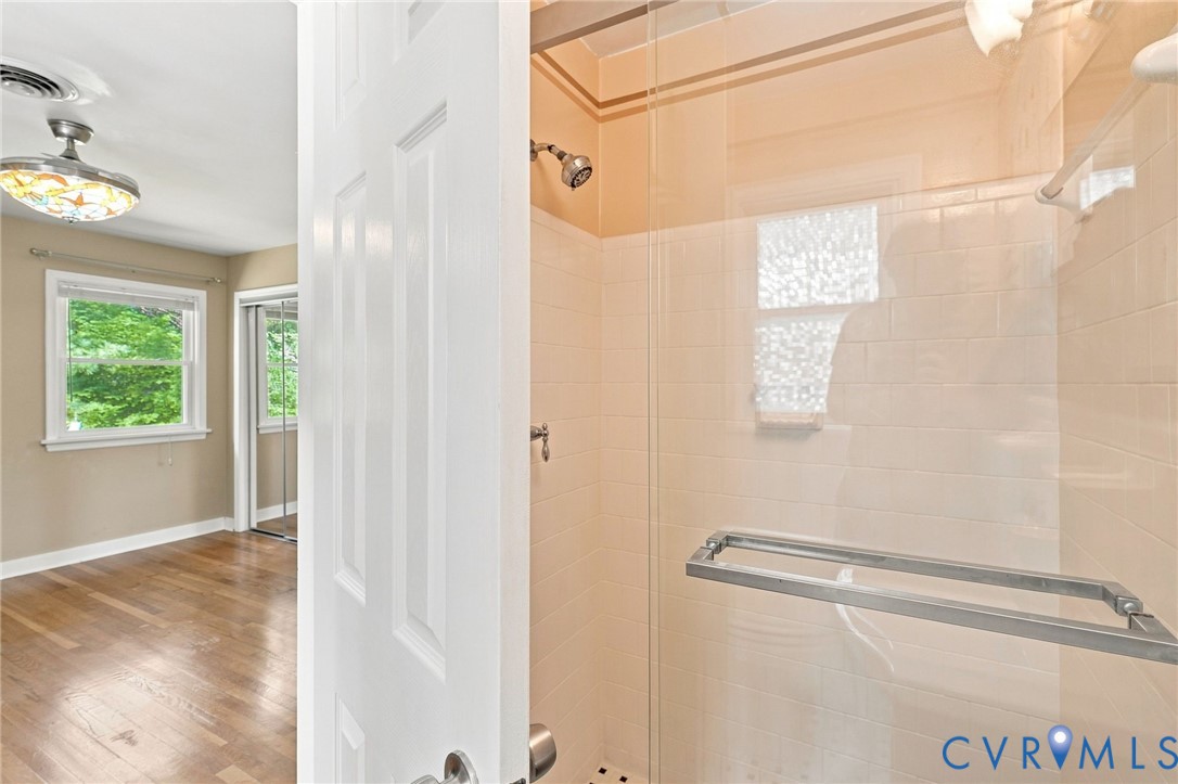 Tiled shower with glass enclosure and chrome fixtures, viewed through a doorway beside a hardwood-floored room.