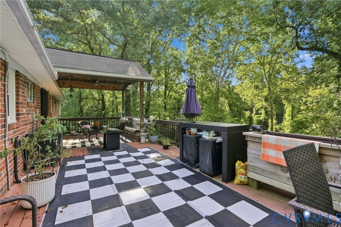 Wooden deck with a checkerboard rug, outdoor seating, potted plants, and a covered barbecue.