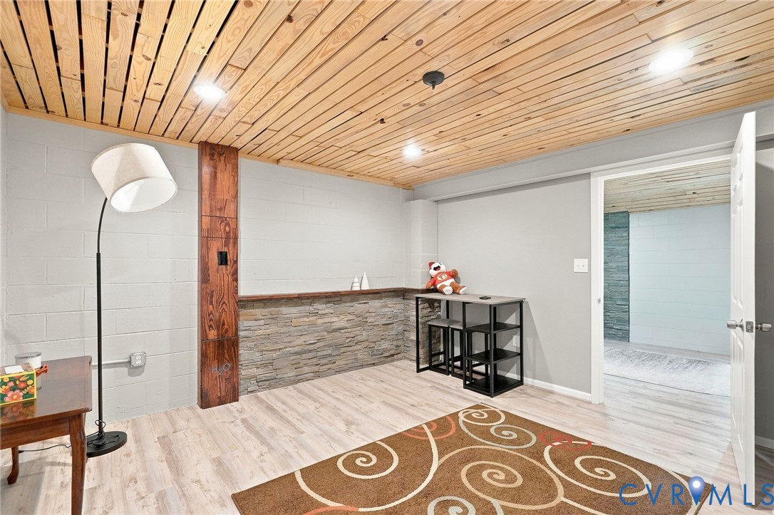 Finished room with a wood plank ceiling, stone-faced half wall, and open doorway to an adjoining room.