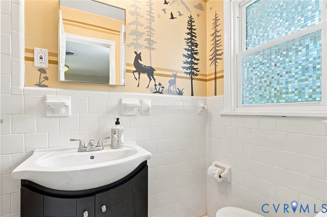 Bathroom with a dark vanity, white tile, blue mosaic window, and woodland mural.
