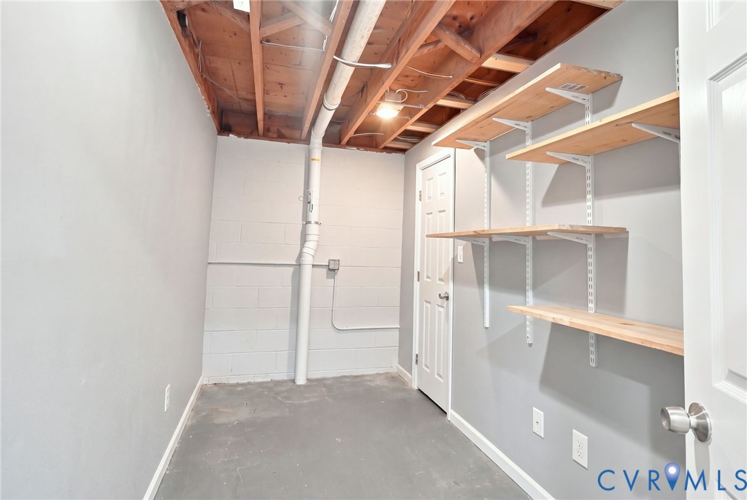Basement storage room with exposed ceiling joists, a utility pipe, and wall-mounted shelves.