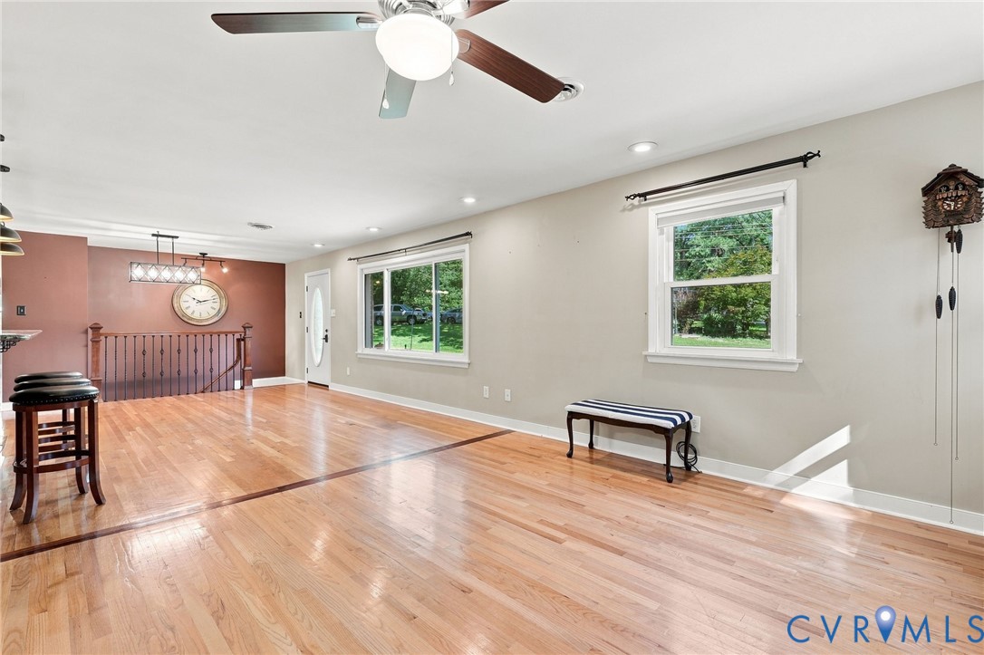Living room with hardwood floors, a ceiling fan, and windows overlooking greenery.