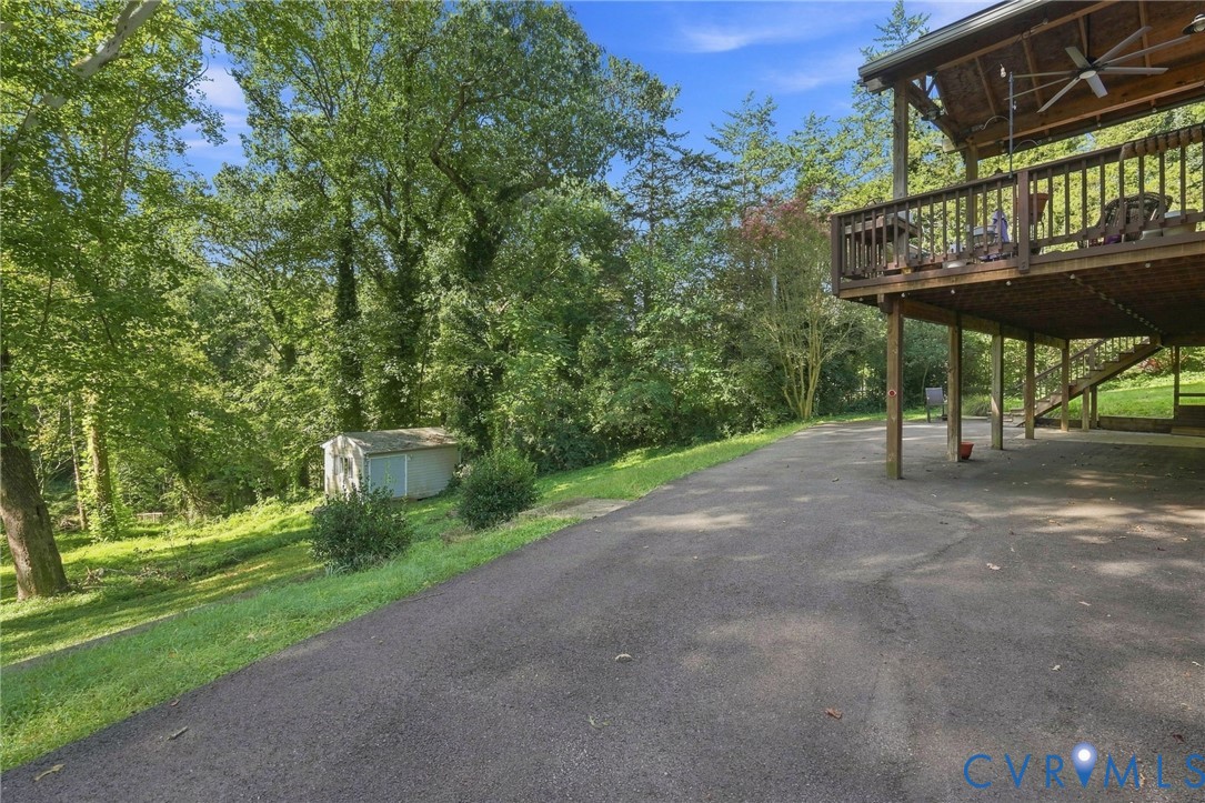 Paved driveway beneath a raised wooden deck, overlooking a wooded yard and small shed.