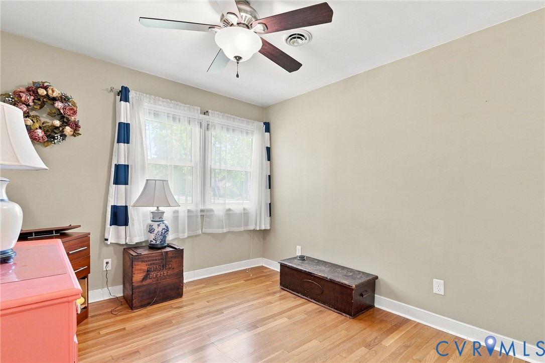 Bright room with hardwood floors, two curtained windows, a ceiling fan, and wooden storage chests.