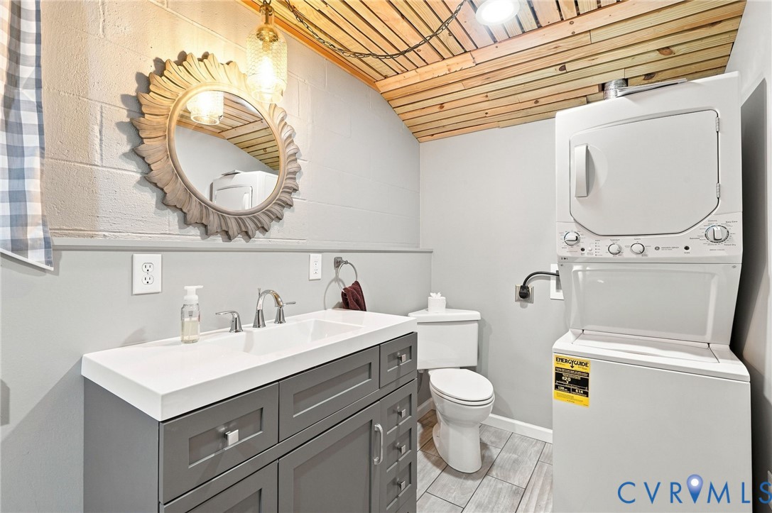 Bathroom with gray vanity, toilet, stacked washer and dryer, and a wood-plank ceiling.