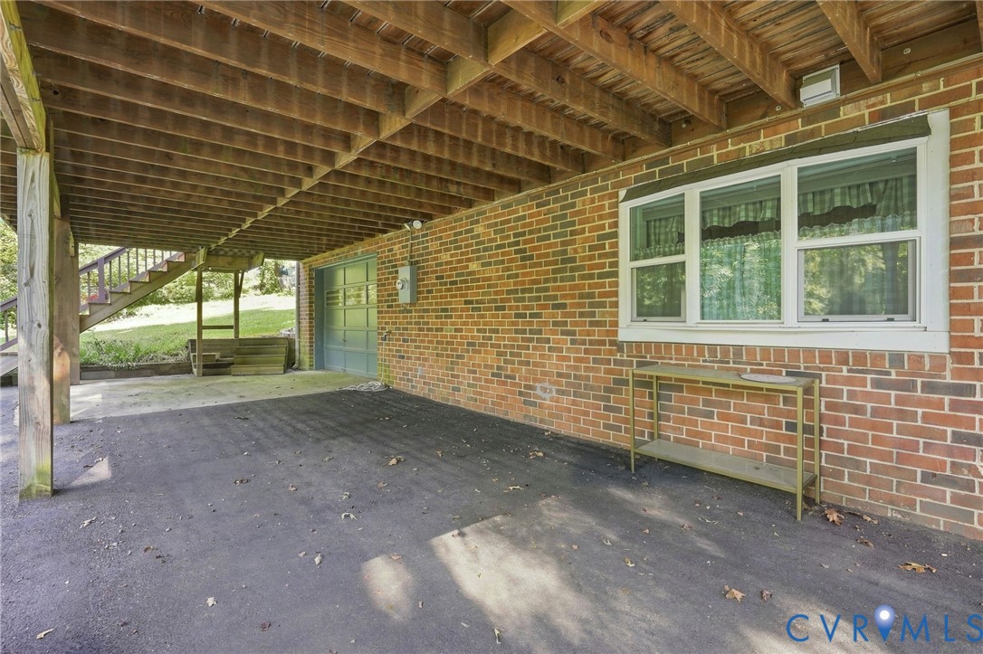 Covered carport beneath a raised wooden deck, beside a brick wall with a garage door and window.