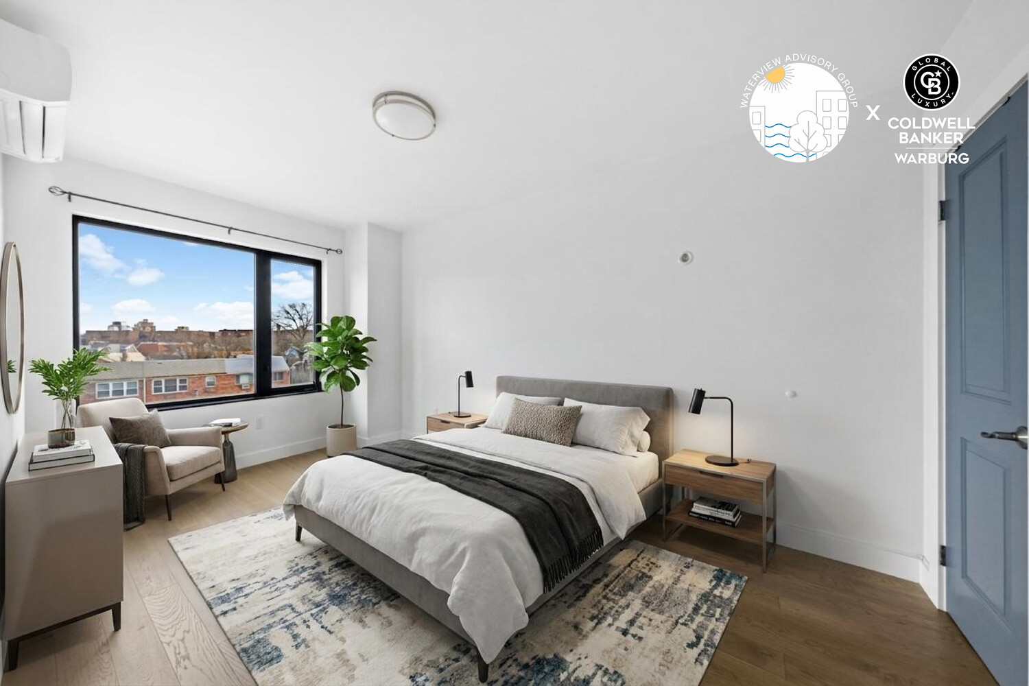 Staged bedroom with a large window, a neatly made bed, and a small seating area.