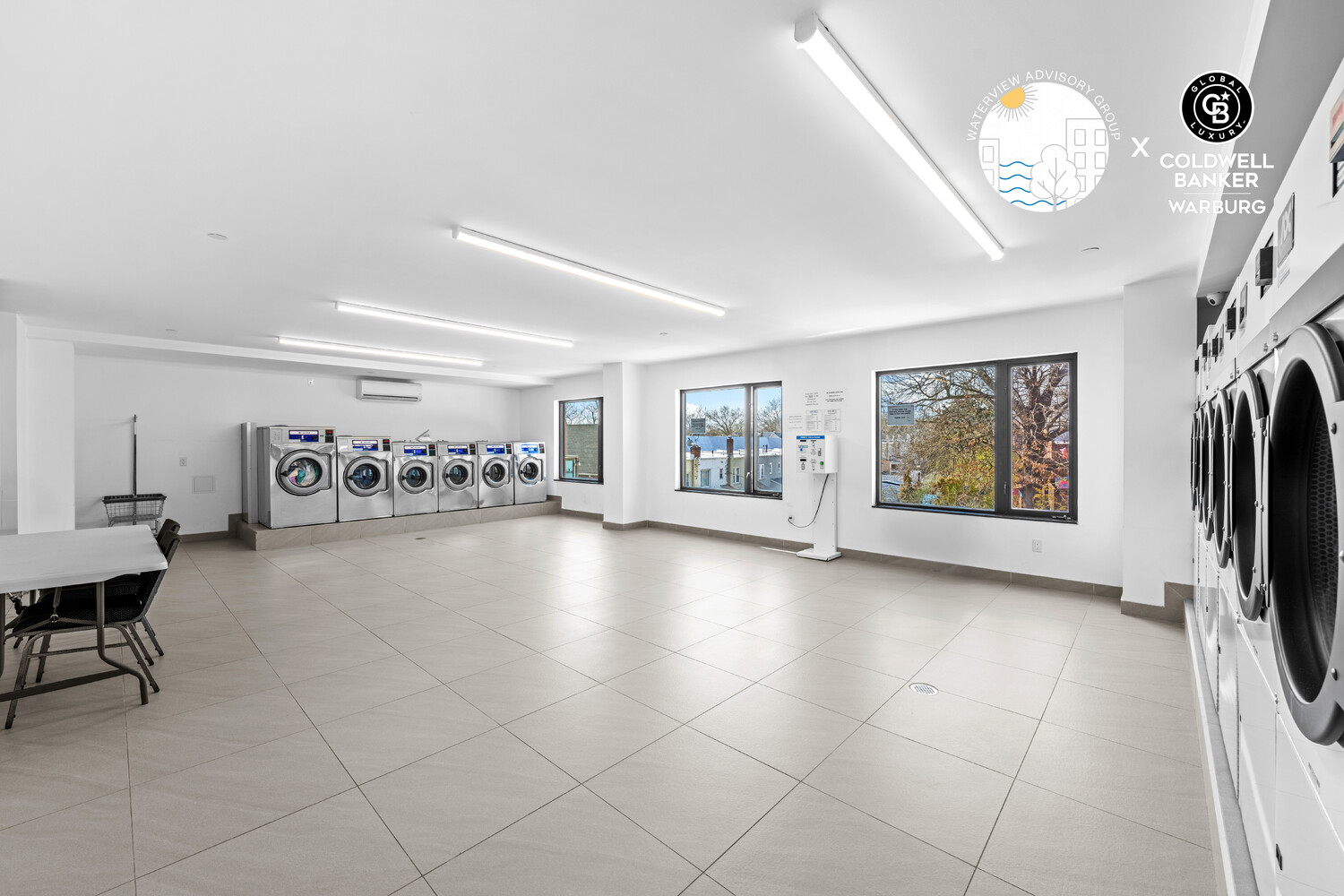 Shared laundry room with rows of front-loading machines, large windows, and a tiled floor.