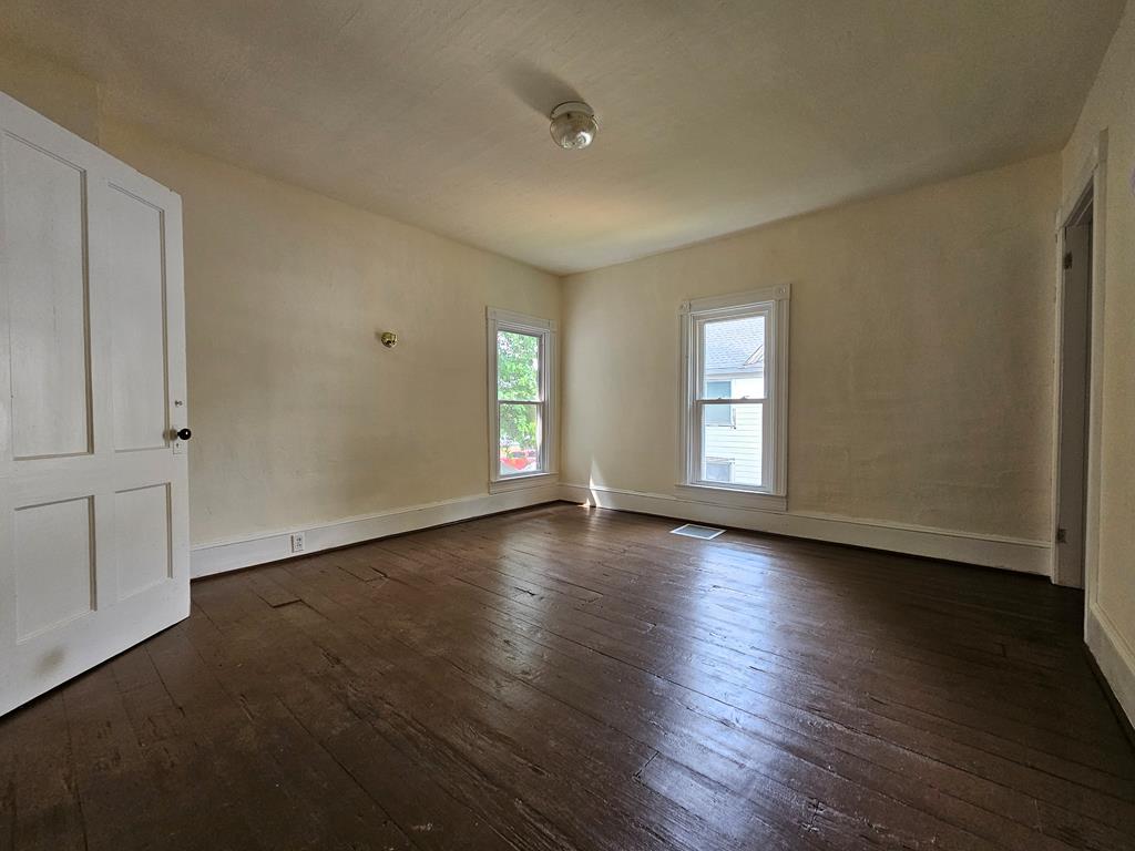 Empty room with worn dark hardwood floors, beige walls, and two windows.