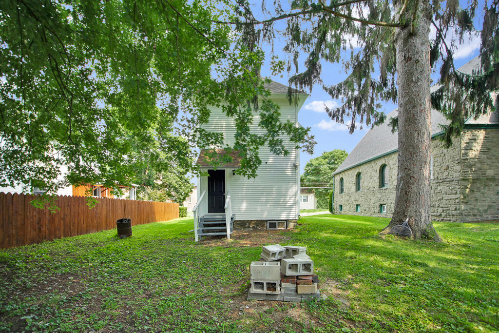 Small white-sided house with entry steps in a shaded grassy yard beside a stone building.