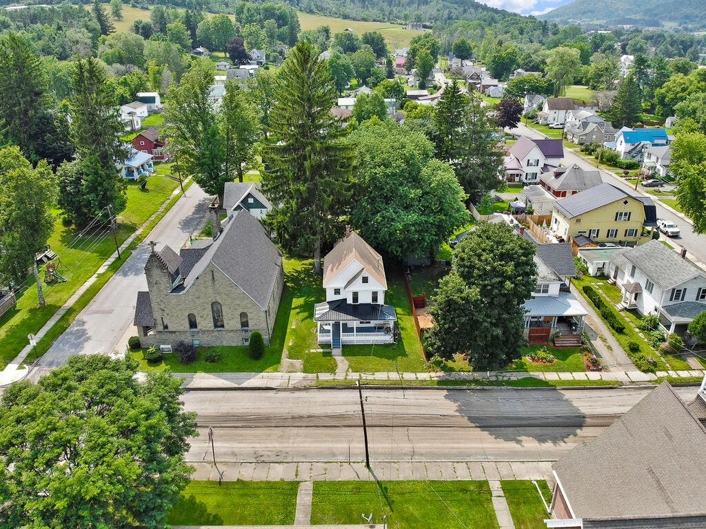 White house with a front porch beside a stone church, viewed from above in a leafy town.