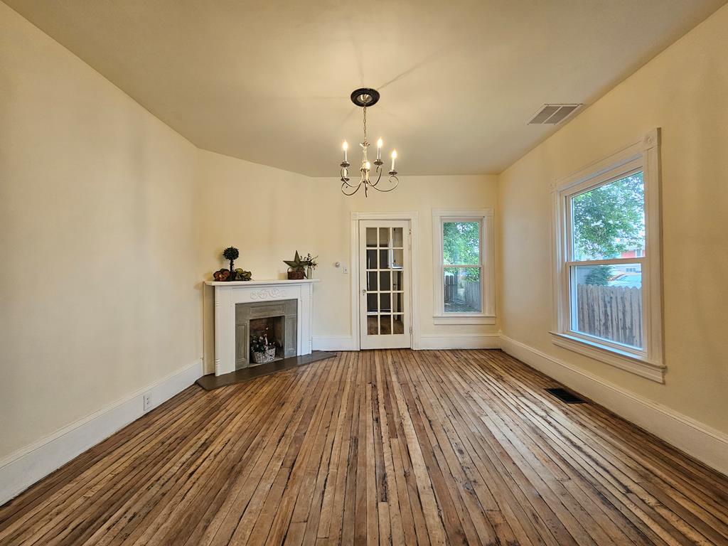 Empty room with a fireplace, hardwood floors, chandelier, and tall windows.