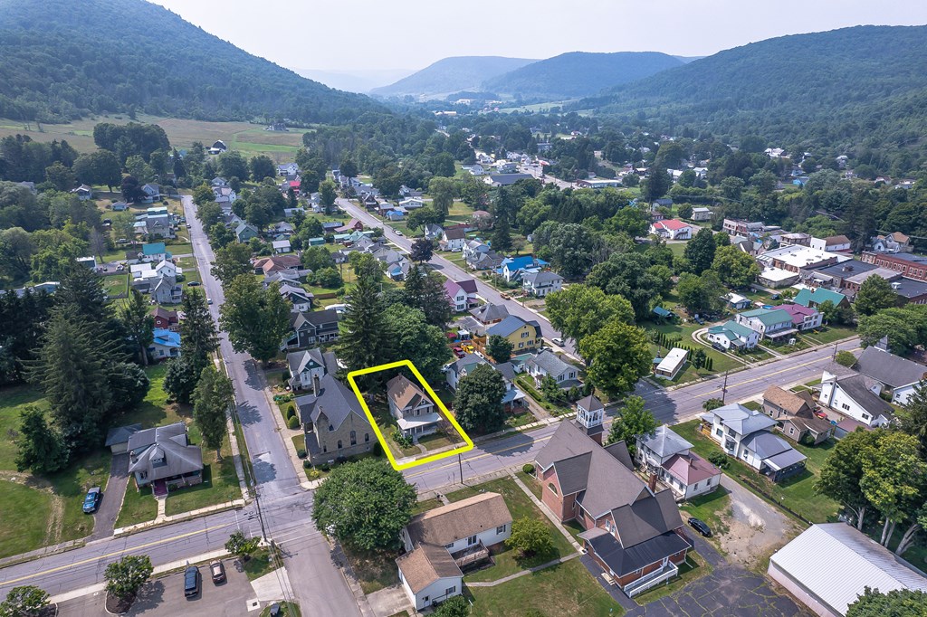 Aerial view of a yellow-outlined home along a town street, with wooded hills beyond.