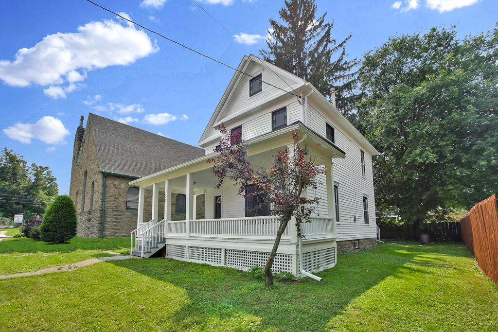 White two-story house with a wraparound porch beside a stone building.