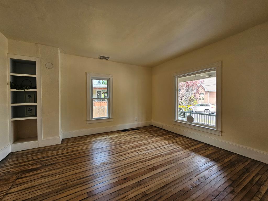 Empty living room with hardwood floors, built-in shelving, and two street-facing windows.