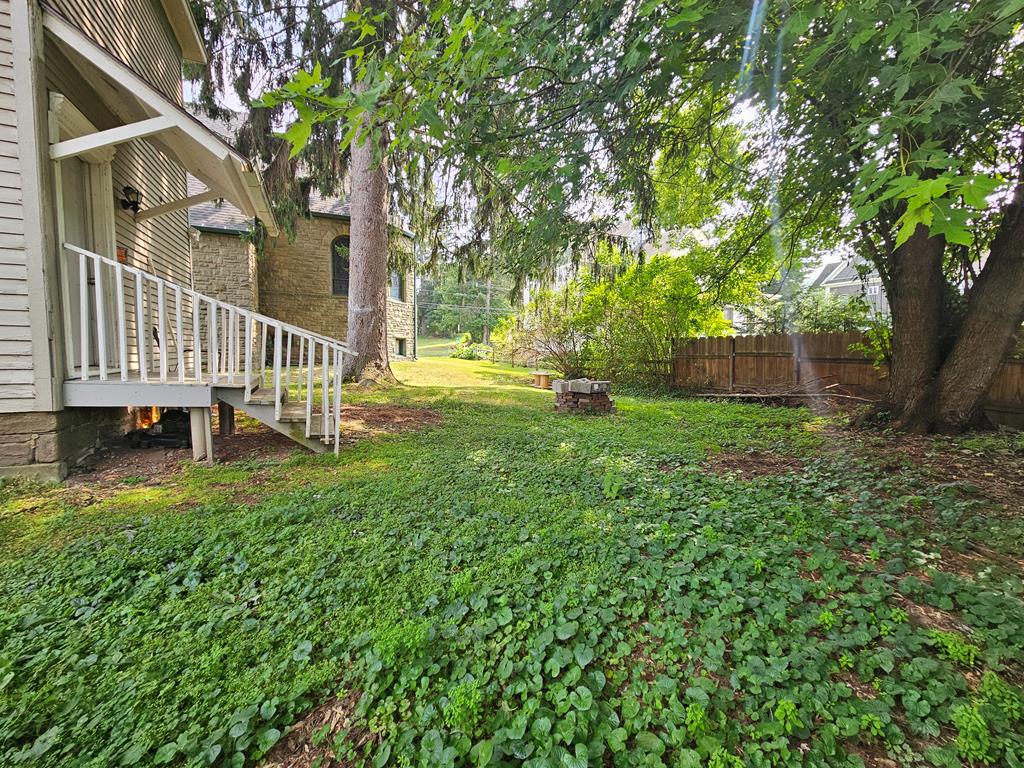 Shaded backyard with a raised porch, mature trees, groundcover, and a small brick fire pit.
