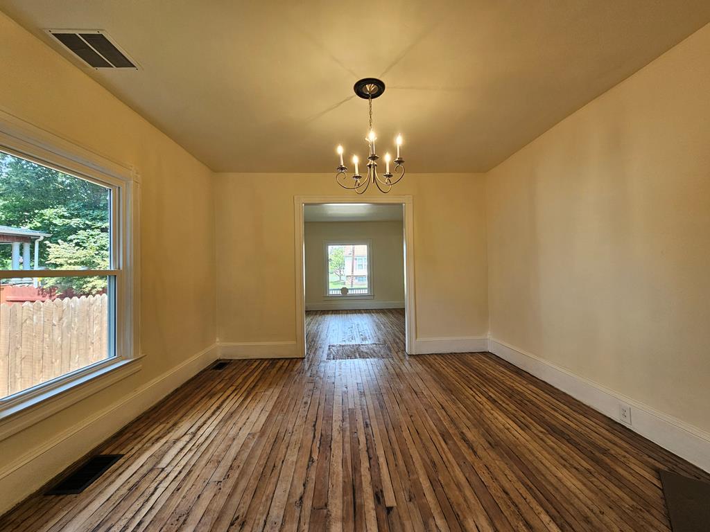 Vacant room with worn hardwood floors, a chandelier, and a large window overlooking a fenced yard.