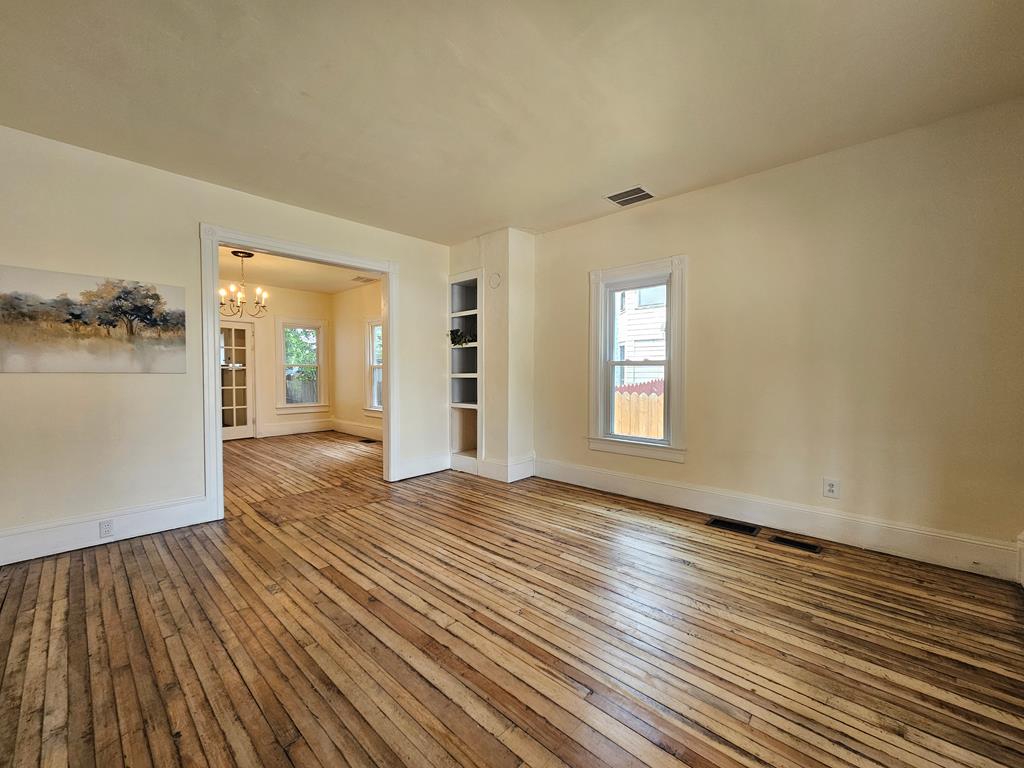 Empty room with wide-plank hardwood floors, built-in shelves, and a doorway to a chandelier-lit room.