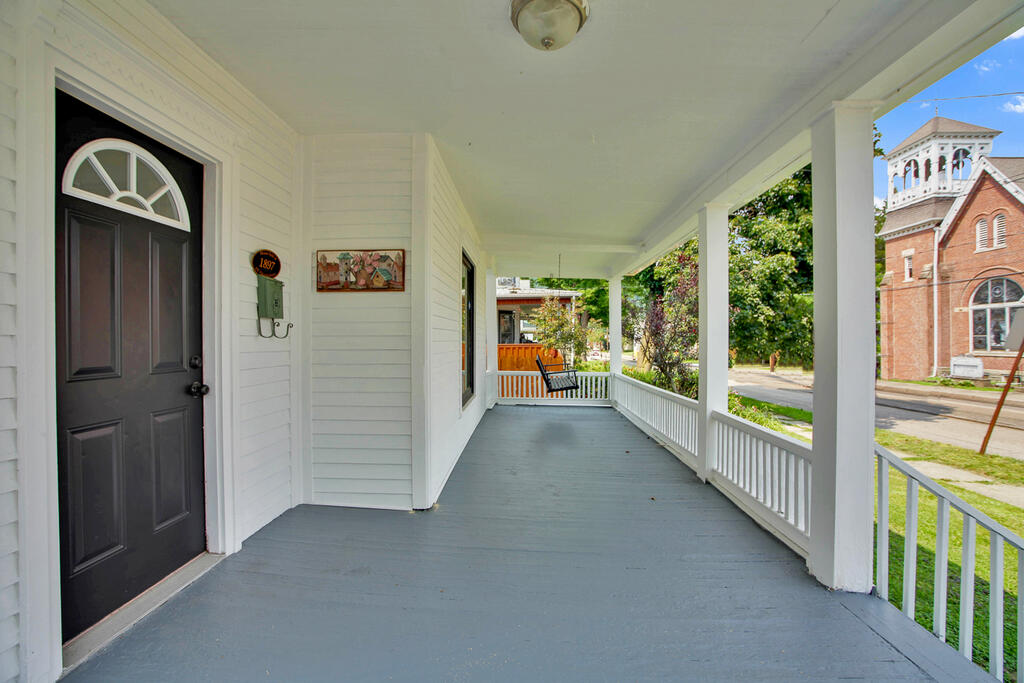 Covered front porch with gray flooring, white railings, and a black entry door; a brick church stands across the street.