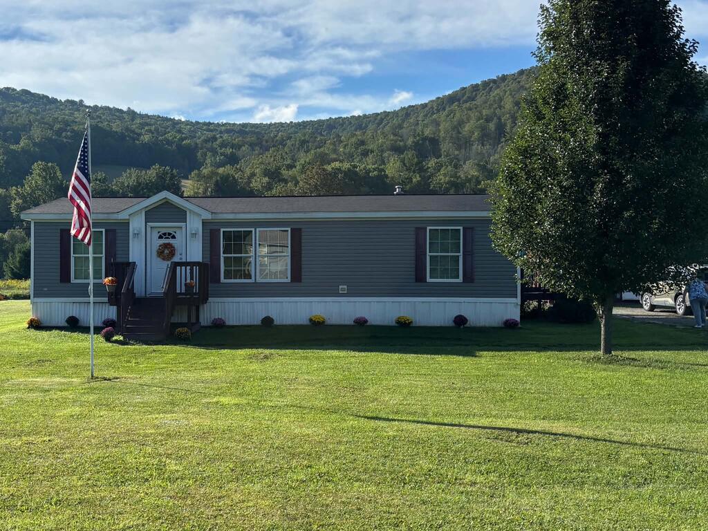 Single-story manufactured home with an American flag, broad lawn, and wooded hills behind it.