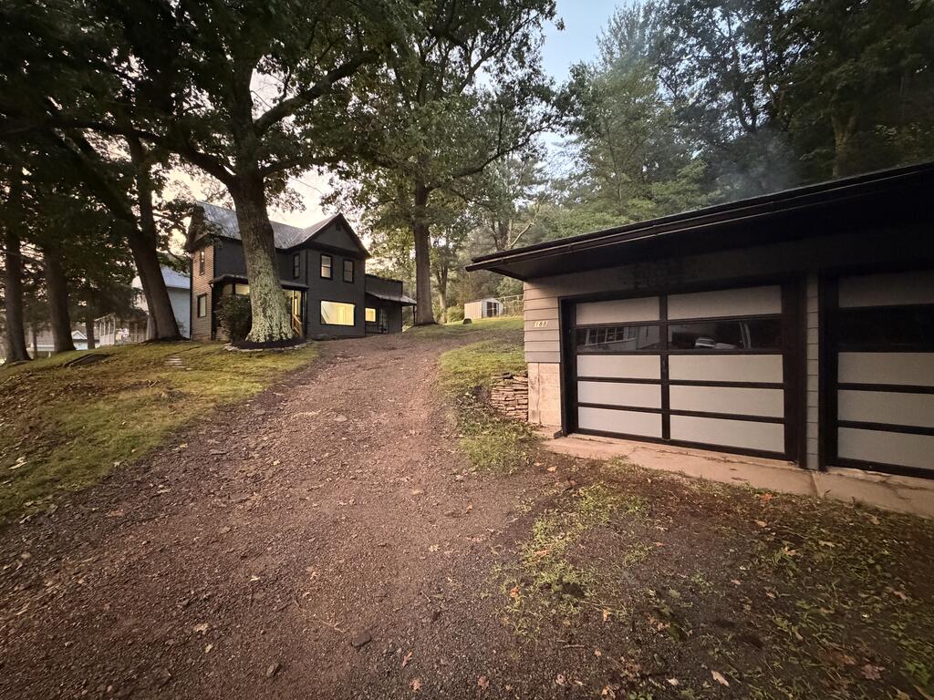 Dark two-story house among mature trees, viewed up a gravel driveway beside a detached garage.