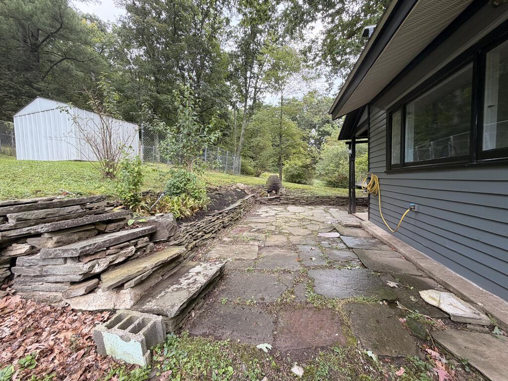 Stone patio beside a gray-sided house, with stacked-stone walls and a white shed uphill.
