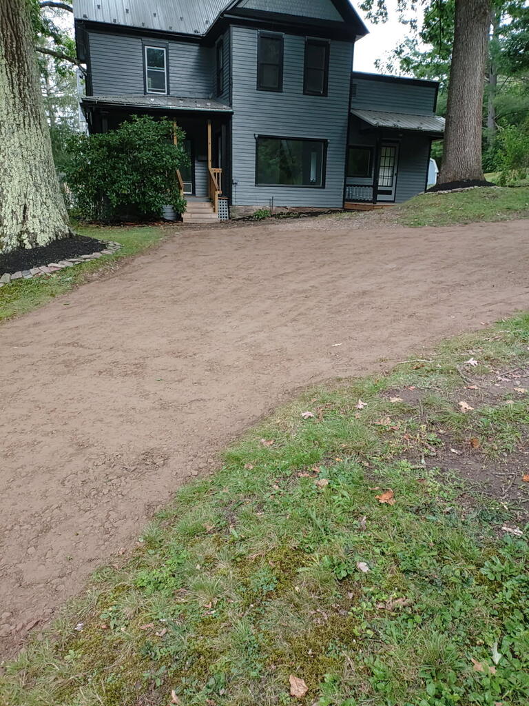 Two-story gray house with a metal roof, mature trees, and a dirt driveway.