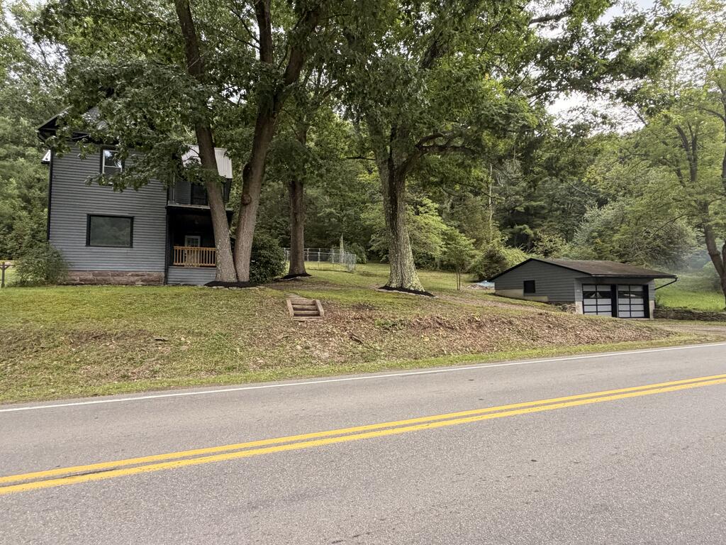 Gray multi-level house and detached garage among mature trees beside a road.