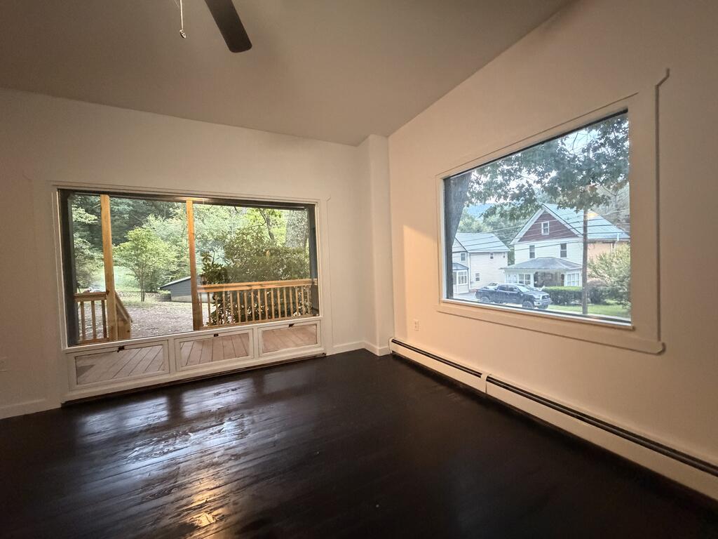 Empty room with dark hardwood floors, large windows, and views of a porch and neighboring houses.
