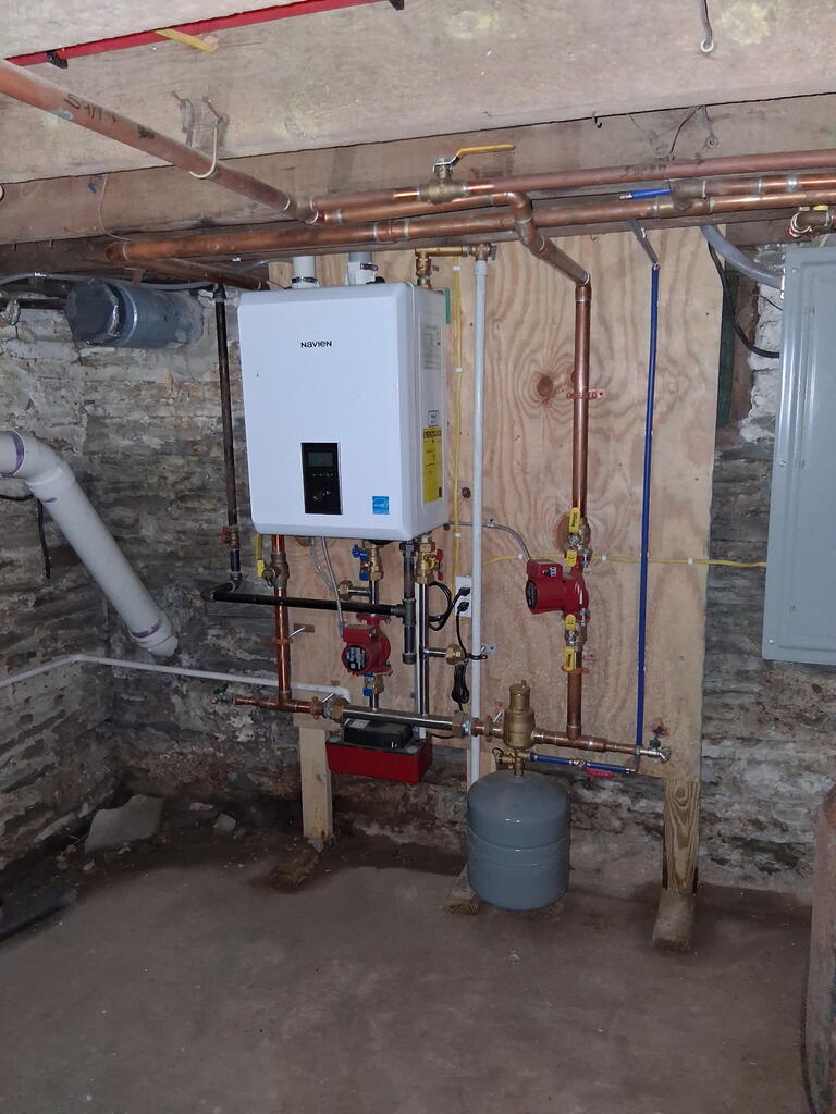 Basement mechanical room with a Navien boiler and exposed copper pipes against stone walls.