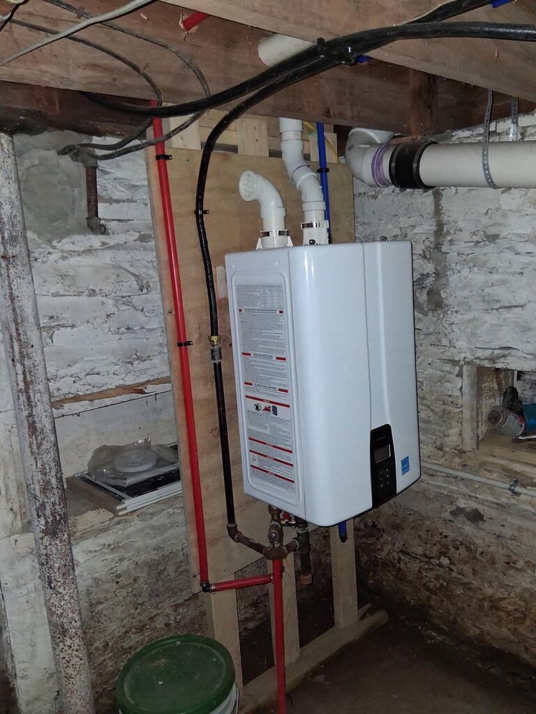 Basement tankless water heater mounted against rough stone foundation walls, with exposed plumbing and ceiling joists.