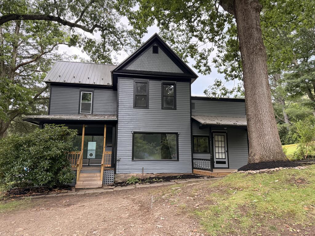 Gray two-story house with a steep gable, covered porch, and large tree beside the entrance.