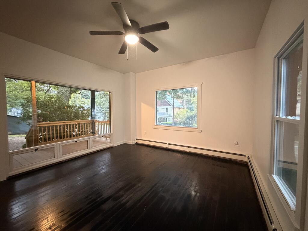 Empty room with dark hardwood floors, a ceiling fan, and large windows overlooking a wooden deck.