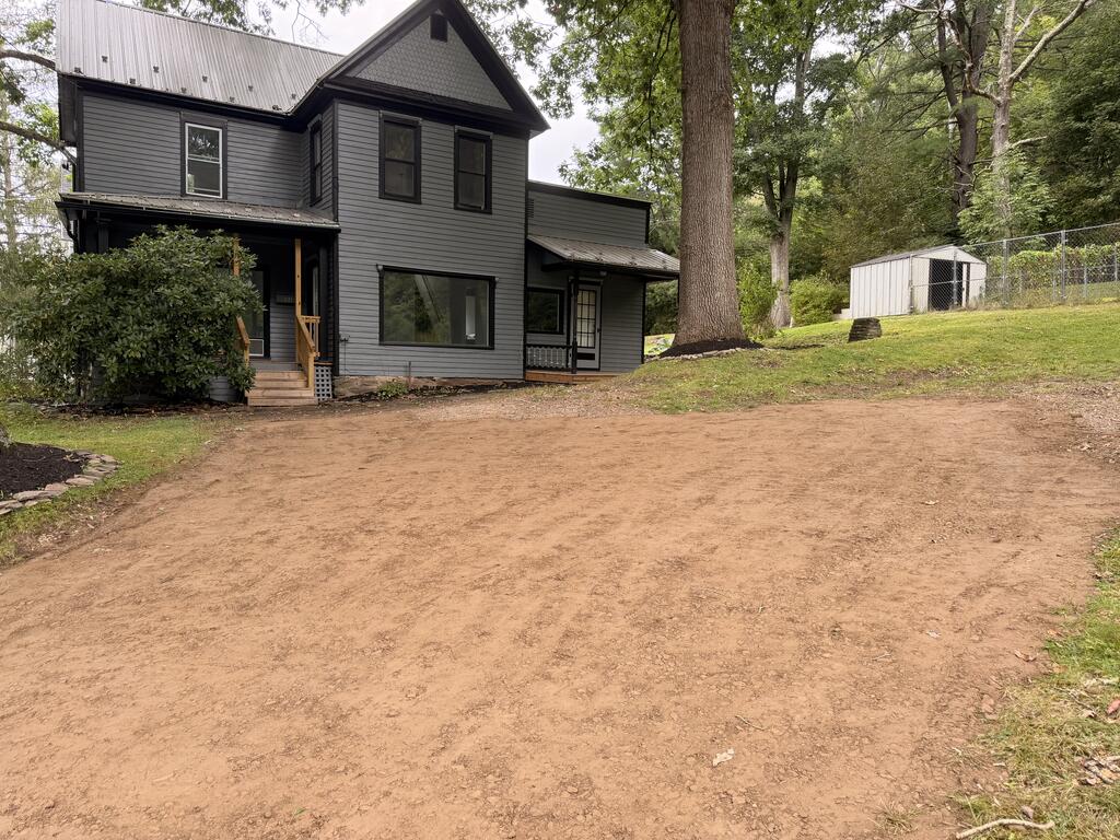 Dark gray two-story house with a covered porch beside a broad dirt driveway and mature trees.