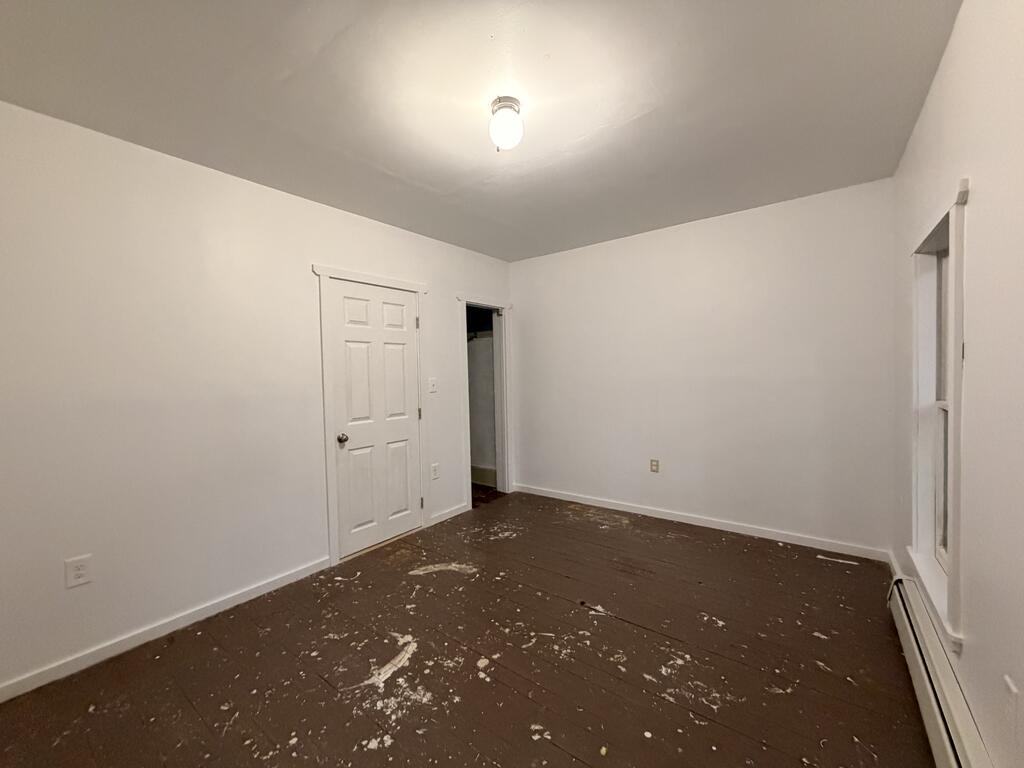Vacant room with white walls, worn dark plank flooring, and a window.