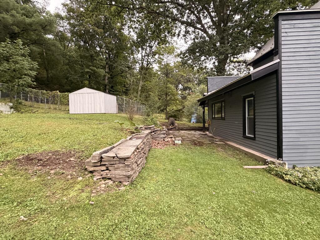 Side yard with a stone retaining wall, white shed, and mature trees beside a gray house.