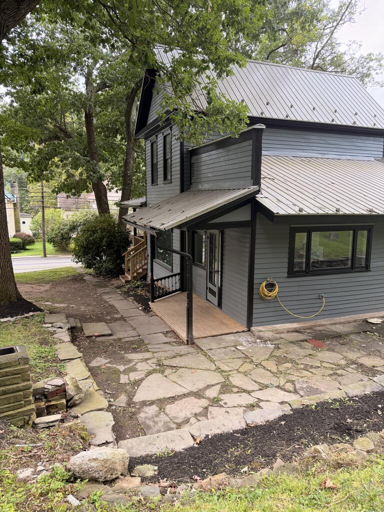 Gray two-story house with a metal roof, covered porch, and stone walkway beneath mature trees.
