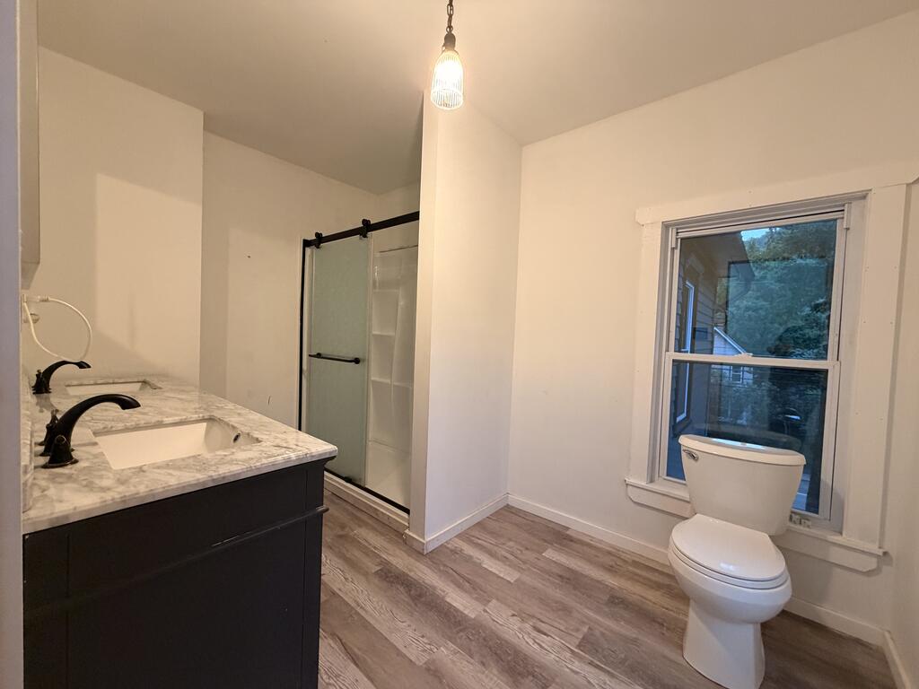 Bathroom with a dark double vanity, glass shower, and toilet beside a window.
