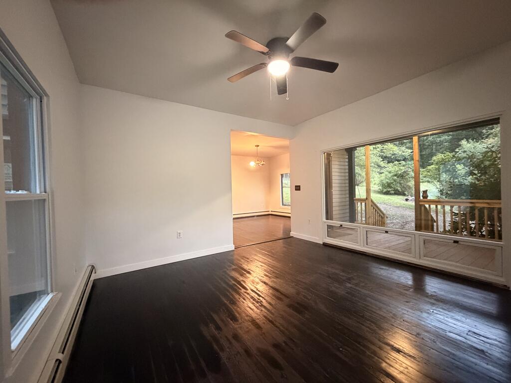 Empty living room with dark hardwood floors, ceiling fan, and large windows overlooking trees.