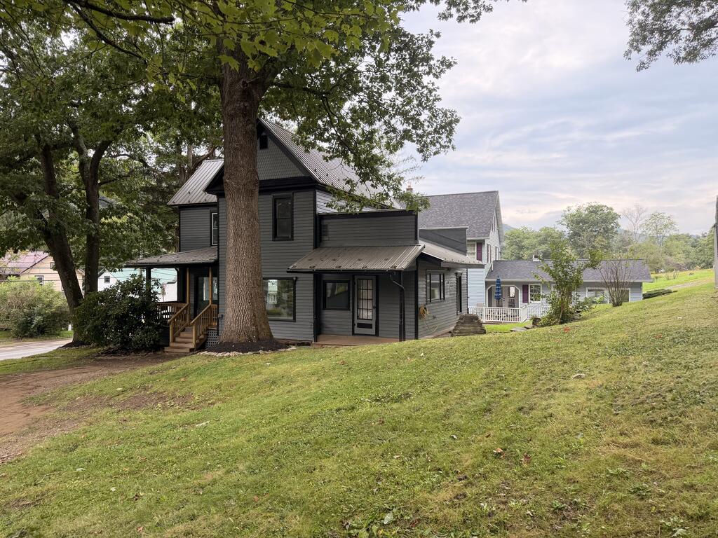 Dark gray two-story house with a steep gabled roof and porch beneath large shade trees.