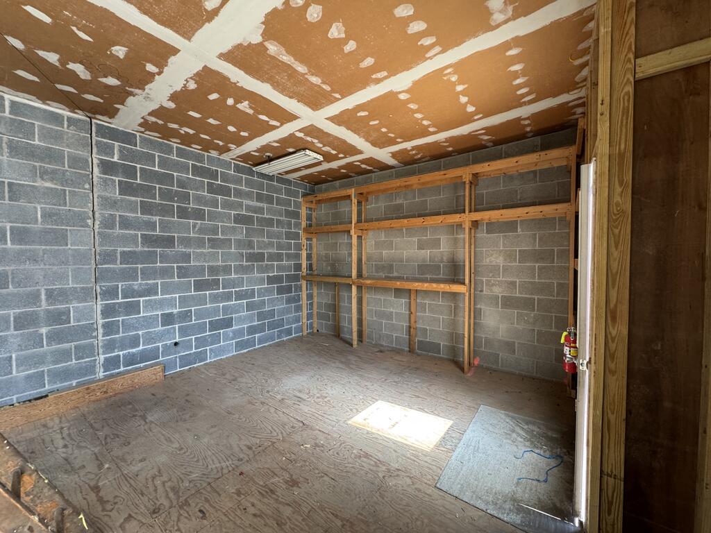 Unfinished garage with concrete block walls, wooden shelving, and a plywood floor.