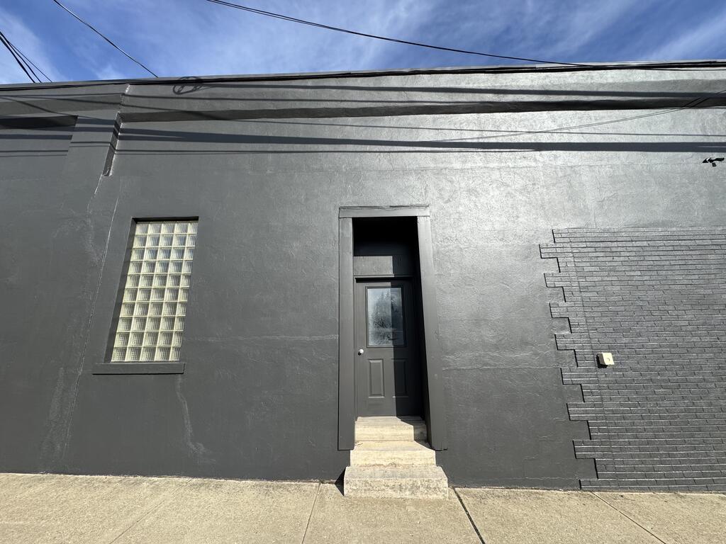 Dark gray building facade with a recessed entry, concrete steps, and a glass-block window.