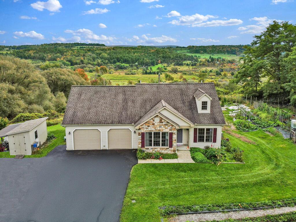Aerial view of a house with a stone entry, two-car garage, and wooded countryside beyond.